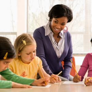 A woman smiling at three young girls who are writing.