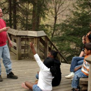 A man is talking to a group of children outside and they are raising their hands.