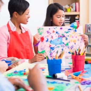 A teacher and her students at an art table and one student is showing off her painting.