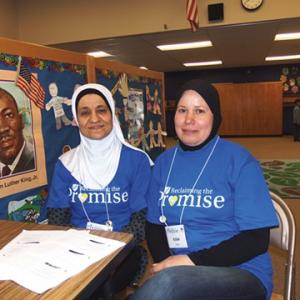Two women in hijab sitting at a table and smiling at the camera.