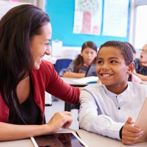 A teacher and student smile at each other while the boy holds a tablet computer.