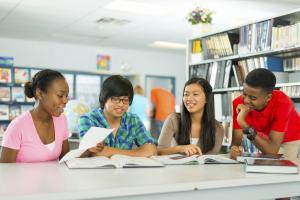 four smiling young adults sitting at table that has books on it