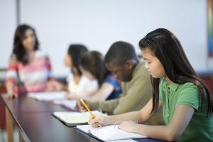 A woman and four children sit at a table and the children are writing