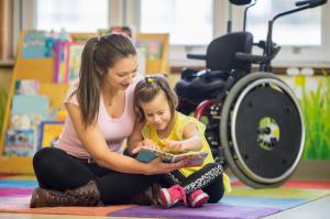 A woman sitting on the floor reading with a child and there is a wheelchair behind them.