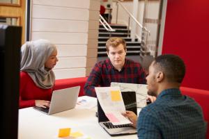 three adults with laptops talking at a table. the woman wears a hijab.