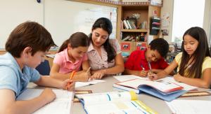 four children writing at a table. a woman helps one of them.