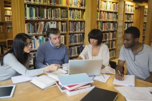 four adults sitting around a table covered in books and looking at a laptop