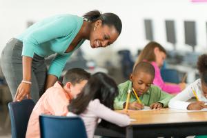 three children writing at a table while a smiling woman leans over them