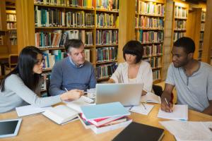 four adults sitting around a table covered in books and looking at a laptop