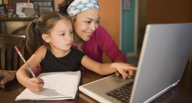 A woman and child are looking at a laptop screen and the girl is writing