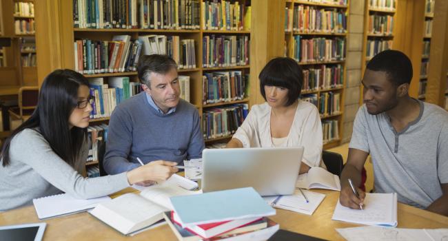 four adults sitting around a table covered in books and looking at a laptop